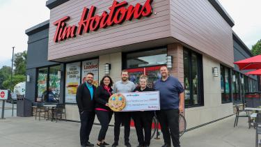 Owners of Tim Hortons stand in front of Tim Hortons holding large cheque