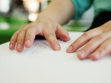 Close-up hands of person with blindness disability using fingers reading Braille book studying in library.