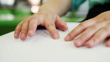 Close-up hands of person with blindness disability using fingers reading Braille book studying in library.