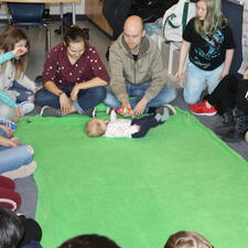 Group of students students sit around a large circle and watch the dad and baby visitors